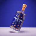 A bottle of Pilgrims Original Gin leaning on its side against a blue studio background, with a white surface beneath it and a visible brand label.