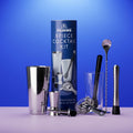 Pilrims 8-piece cocktail kit displayed with its blue cylindrical packaging box on a light purple surface against a blue gradient background. The set includes a stainless steel shaker, mixing glass, strainer, bar spoon, muddler, jigger, and other bar tools.