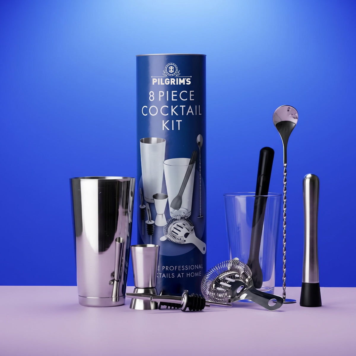 Pilrims 8-piece cocktail kit displayed with its blue cylindrical packaging box on a light purple surface against a blue gradient background. The set includes a stainless steel shaker, mixing glass, strainer, bar spoon, muddler, jigger, and other bar tools.